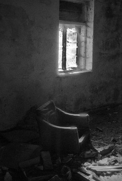 The black and white image depicts a dimly lit, abandoned room. A small window allows a shaft of light to enter, casting harsh illumination on the interior. In the foreground, there is an old, worn-out armchair, partly lit by the window’s light. The floor is cluttered with debris, broken wood, and scattered materials, indicating long-term neglect and decay. The rough texture of the walls and the general darkness contribute to a somber, melancholic atmosphere.