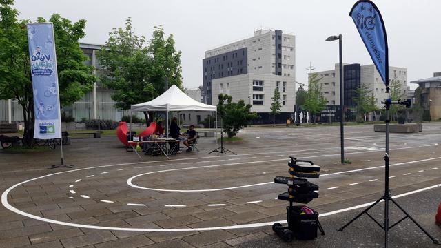 Une piste vélo-école tracée sur une grande place dégagée, avec l'oriflamme Vélodays, un barnum sous lequel s'abrite un groupe de personnes. Et au premier plan l'oriflamme d'Hérouelibre et une servante à outils ouverte.