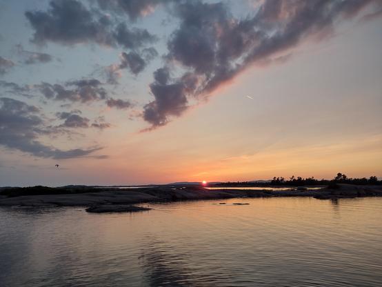 Purple and red sunset looking out to the horizon over calm water with numerous low, smooth rock islands and some pine trees on the shore in the far distance. Purple clouds float in a dimming blue sky.