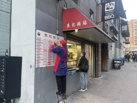 A woman who works at the small snacks shop (dumplings, etc) stands on a small stool to update prices on the big board. More expensive (in case this is unclear). 41st Ave
