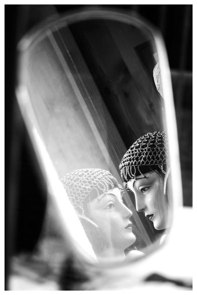 Schwarz-Weiß-Foto im Hochformat mit weißem Rand. Im Schaufenster eines Frisiersalons steht ein ovaler Spiegel. In diesem spiegelt sich der Kopf einer Schaufensterpuppe zweimal, dadurch sieht es so aus, als ob die Puppe sich selbst anschaut. Sie trägt eine Kopfbedeckung, die an die Mode in den 1920ern erinnert.