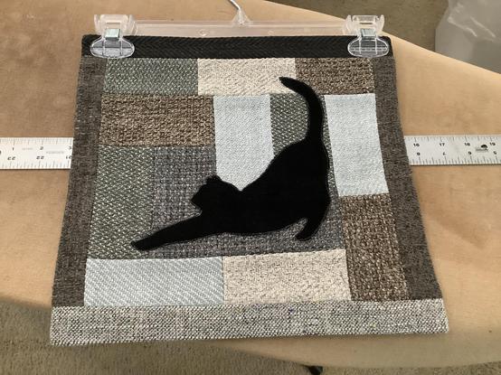 12.25” wide x 11.75” high pieced wall hanging made with rectangles of brown, gray, and pale blue upholstery fabrics. The middle is filled with the black silhouette of a cat stretching with it’s rear in the air and it’s front near the “floor”.