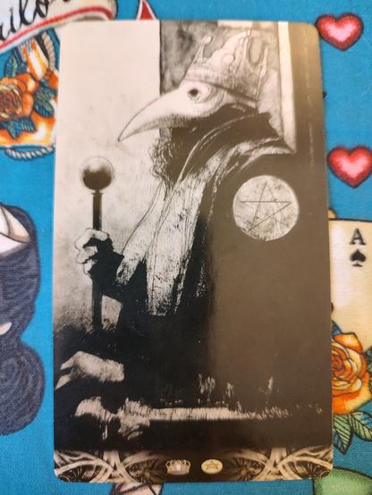 Card from the Murder of Crows Tarot deck. 
King of Pentacles. 
A man wearing a crow's mask, his beard peeking out a bit from under the mask, seated. He holds a scepter on his right hand, and there is a pentacle on his left shoulder. He sits sideways, his left to the viewer.