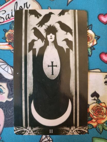 Card from the Murder of Crows Tarot deck.
The High Priestess-II.
A woman dressed in black and white, a black cross in center of her dress. She stands over a crescent moon. Some crows fly above her.