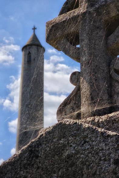 A close-up of a weathered stone Celtic cross, partially covered in spiderwebs, with a tall, round tower featuring a cross at its peak visible in the blurred background under a blue sky with scattered clouds.