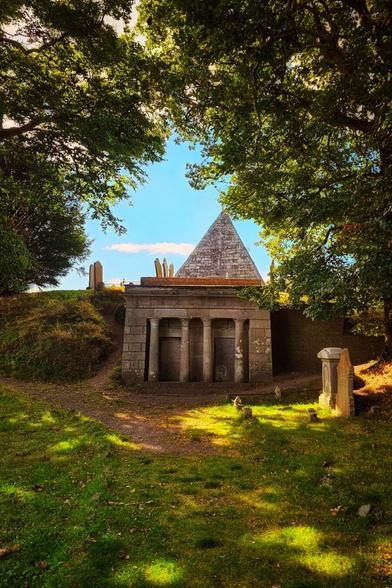 A stone pyramid-shaped mausoleum with a portico stands on a grassy hill, framed by lush green trees under a bright blue sky. Several weathered headstones are visible on the hillside.