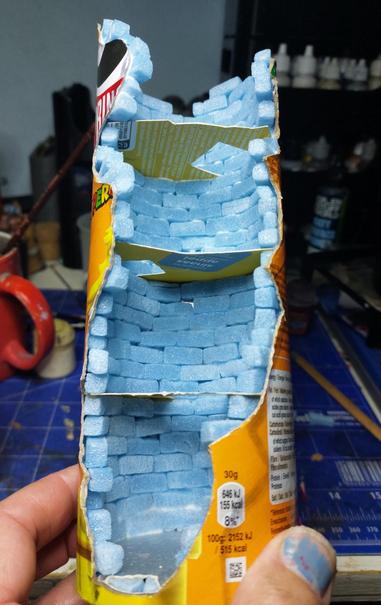 Pringles can with an irregular pattern cut out from one side and small blue xps bricks stuck to it and three extra floors stuck in with pieces of an old tea box.