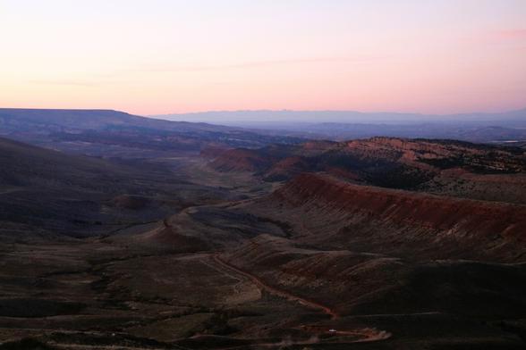Wide-angle view of a rugged canyon landscape at dusk, with layered red rock formations and rolling hills fading into the distance. The sky is softly lit in pastel hues of pink and lavender, casting a tranquil glow over the terrain. A dusty dirt road winds through the foreground, with a small vehicle kicking up dust near the bottom of the image.