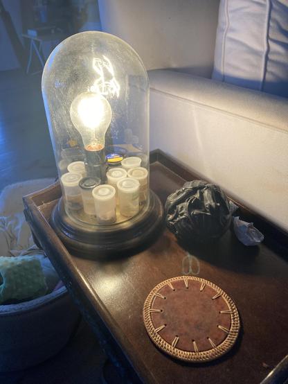 Glass domed lamp over naked bulb and plastic film canisters filled with water from around the world. Sitting on a wooden side table with a  woven bamboo coaster and a black ball of carved dragons.