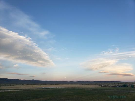 Broad green valley with a nearly full moon rising over low brown hills on the Eastern horizon. Dusk sky with two sets of lenticular clouds. Photo by Jenifer Hanen.