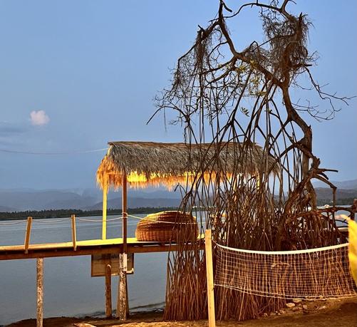 A scenic view of a dock with a thatched-roof structure at dusk, surrounded by mangrove trees. The water is calm, reflecting the soft evening light, while a volleyball net borders the area.