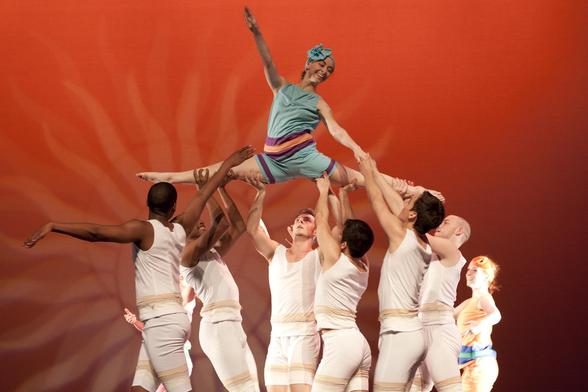 Six male dancers in all white hold aloft a female dancer in a green old fashioned swim suit against a reddish orange background