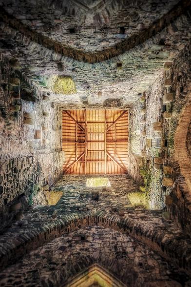 A low-angle photograph looking directly upwards at the interior of Tintern Abbey in Ireland. Weathered, aged stone walls rise vertically, meeting a square wooden roof structure that has been installed to cover the space. The wooden beams and planks of the roof are visible, blocking the sky. The stonework of the abbey walls shows significant signs of age and historical wear.
