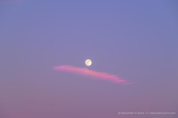 A minimalistic photo showing the full moon centered in the frame, with a pink cloud underneath it. The rest is all empty sky with a gentle gradient from cooler blue to muted pink at the bottom.