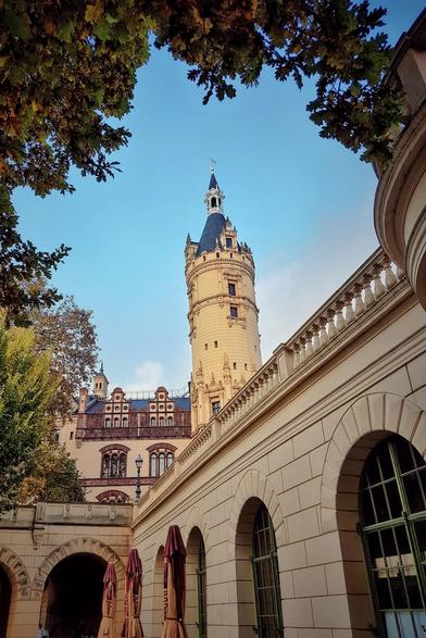 A low-angle shot looking up at a tall, light-coloured turret of Schwerin Castle, framed by autumn leaves in the upper corners. The turret has several small windows and a dark, pointed roof with a spire. To the right, a cream-coloured building with arched windows and a decorative balustrade leads the eye towards the castle. In the foreground, the tops of closed red umbrellas suggest an outdoor seating area. The sky is a clear, pale blue.