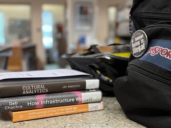 Three library books on a faux marble laminate library circulation desk. A backpack and bicycle helmet sit to the right of the books. Books are titled: Cultural Analytics; How Infrastructure Works; and Anthropological Data in the Digital World