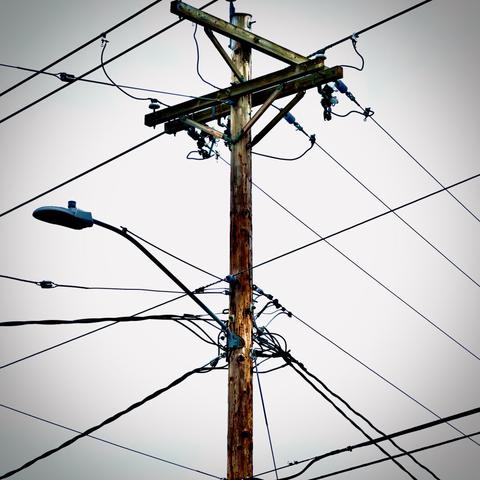 A power pole stands tall, as power lines at different heights and from all four sides extend to it under an overcast sky.