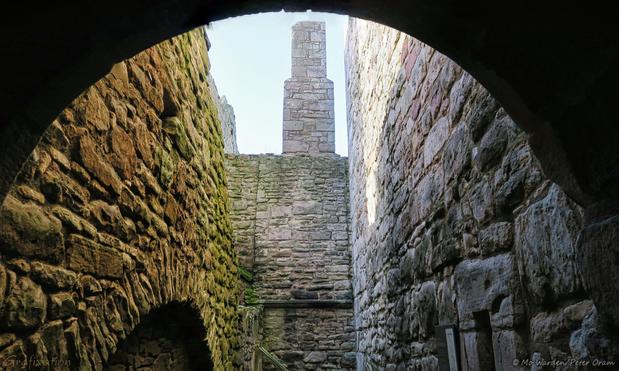 A colour photo taken from inside an archway, looking out at brown and grey brick walls. The stones are ancient and mossy with arches and various sizes of holes in their vertical faces. Ahead is a grey stone wall with a chimney balanced on the top. There is sunlight on the righthand wall and the sky is clear.