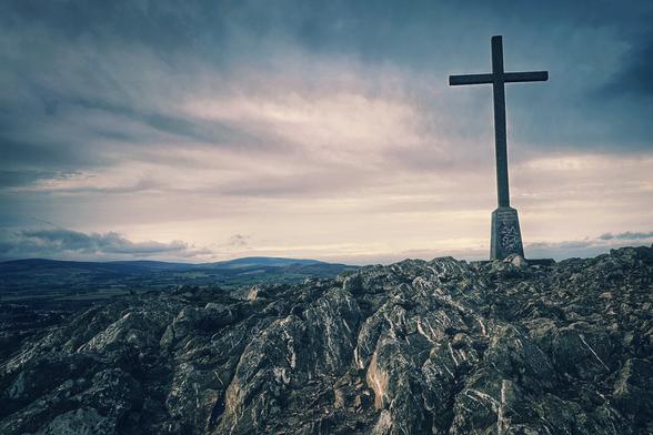 A wide-angle shot shows a large cross atop a rocky hill, with a cloudy sky overhead and a view of the Irish coastline in the distance.