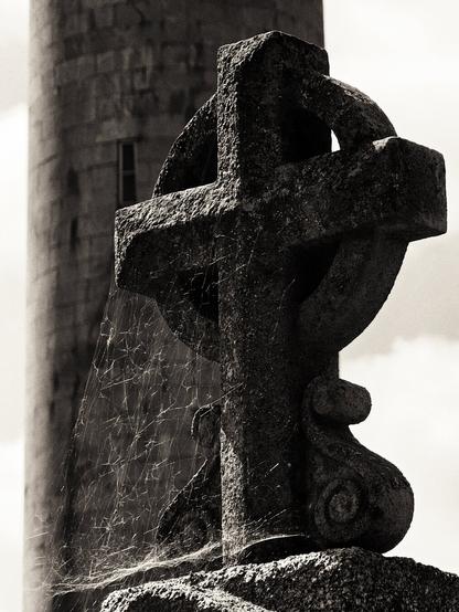 A close-up, sepia-toned photograph shows a weathered stone Celtic cross in the foreground, with a spiderweb draped across its left side. In the blurred background stands a round stone tower under a bright sky.