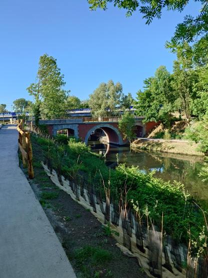 A pedestrian bridge and a water channel in one of the pedestrian paths under construction. Vegetation all around.
Clear sky, sunny weather