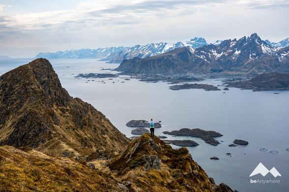A lone runner stands on a rocky summit with arms wide open, gazing out over the island-dotted seascape and jagged, snow-capped peaks of the Lofoten archipelago.