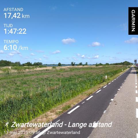 Cyclepath along green pastures with some trees in the background. A small line of water in the middle, blue sky above with a few fluffy white clouds.