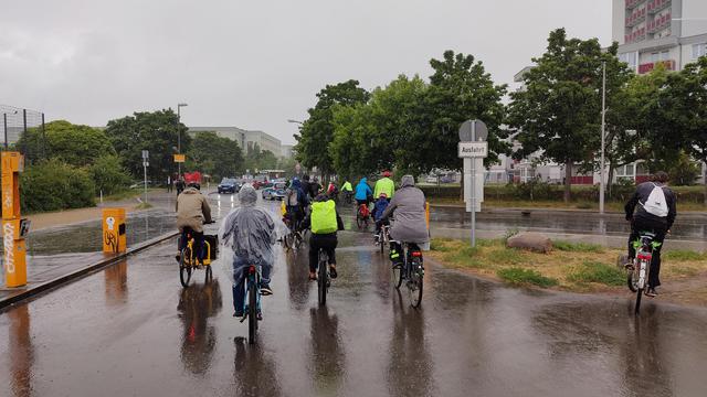Ein Pulk Radfahrender, darunter auch zahlreiche Kinder, fahren von einem Parkplatz auf die Straße. Man kann erkennen, dass es stark regnet.