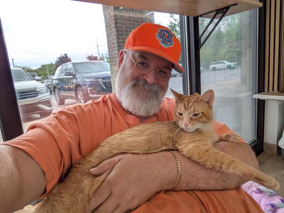 A friendly orange cat relaxing on the chest of a large bearded man in an orange shirt and hat