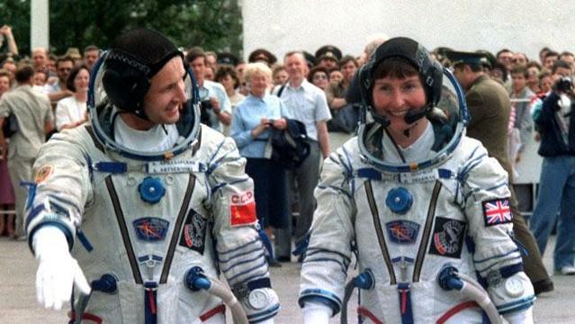 Helen Sharman walking towards the rocket in her spacesuit but carrying her helmet. She's a white woman with dark hair. A male colleague is walking alongside her.