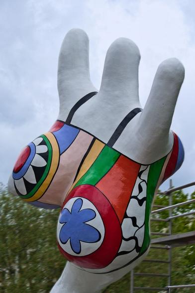 Eine farbenfrohe, abstrakte Skulptur in Form einer Frau mit deutlichen Rundungen, mit bunten Mustern verziert. Die Muster über dem Körper lassen einen an einen Badeanzug denken. Die Skulptur ist von schräg hinten und von unten fotografiert. Es sieht so aus, als würde die Skulptur begeistert und freudig ins Wasser rennen oder hüpfen.

Die Skulptur ist als Hauptmotiv präsent und nimmt den Großteil des Bildes ein. Sie ist mit bunten Formen und Farben bedeckt, die in verschiedenen Größen und Mustern angeordnet sind.  Die Formen erinnern an geometrische Muster, Kreise, Streifen und Blumen. Die Farben sind leuchtend und kontrastierend, wie Rot, Blau, Grün, Gelb und Weiß. 

Die Umgebung ist unscharf und die Beleuchtung ist natürlich und scheint von oben zu kommen. Die Hintergrunddetails sind nur vage erkennbar, und sie geben die Atmosphäre von außen in einem Park wieder.