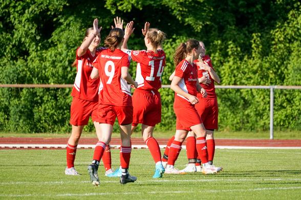 Eine Gruppe von Fußballspielerinnen in roten Trikots feiert gemeinsam auf dem Spielfeld, vermutlich nach einem Tor. Im Hintergrund ist eine grüne Hecke zu sehen.