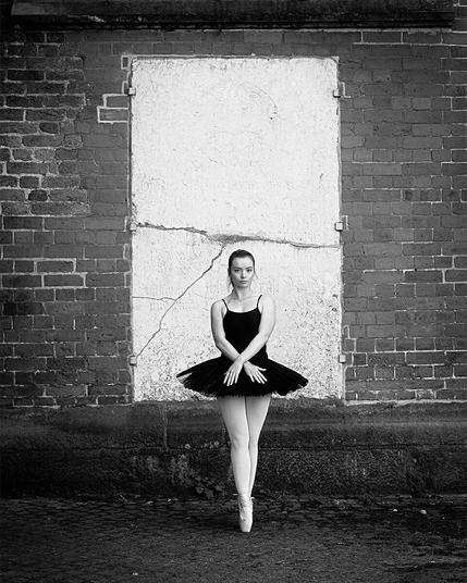Ballet dancer standing on lace in front of an old church wall. Her black tutu stands out against the white tombstone embedded in the wall.