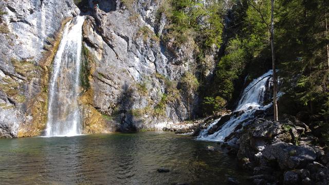 Zwei Wasserfälle, die sich in ein Wasserbecken ergießen. Der linke Wasserfall fällt senkrecht ab, während der rechte Wasserfall über Steine ins Wasserbecken rinnt.
