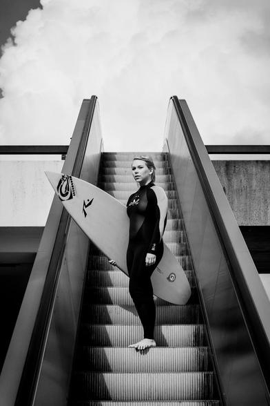 Surferin im Neo mit Surfbrett auf Rolltreppe stehend