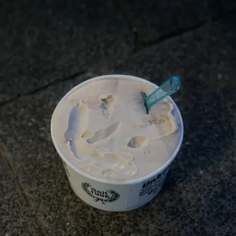 Becher gestrichen voll mit Eis. Ein blauer Eislöffel aus Plastik steckt im Eis. Ein Smiley ist angedeutet.