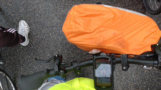 Blick herunter auf meine Regenjacke, Hose, den Lenker und die Fronttasche von meinem Brompton Electric G-Line: Ich trage eine neogelbe Regenjacke, reflektierende Rainlegs über den Oberschenkeln, unter den Rainlegs eine lange Blue Jeans. Über die Fronttasche ist ein teilweise eng anliegender, orangefarbener Regenschutz mit vorne silbernem Aufdruck drüber gezogen.