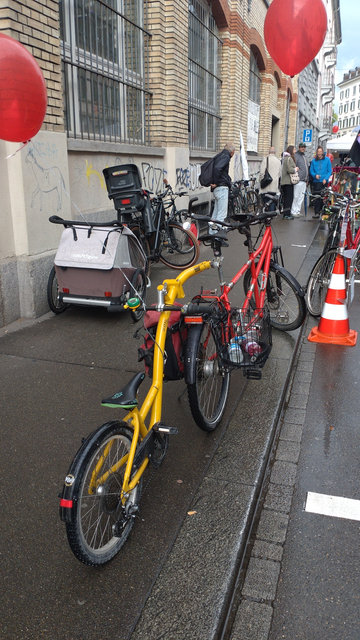 Auf dem Gehweg steht ein rotes Tandem mit einem gelben, halben Kinderrad als Anhänger bzw. Nachläufer. Blick von schräg hinten.
