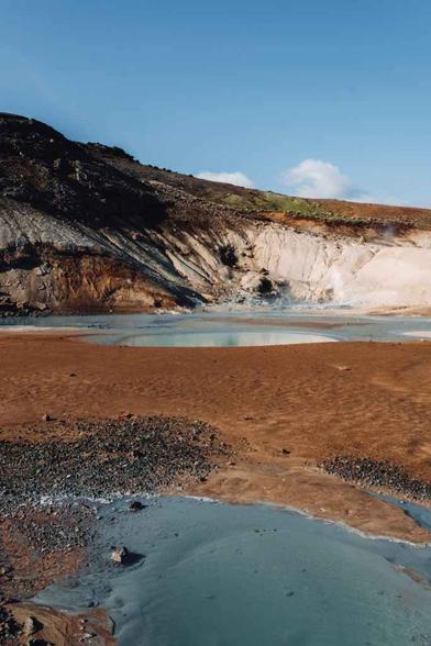 Photo of a hot spring and geyser with a foreground of rough reds, the pool of water and a cliff in the background