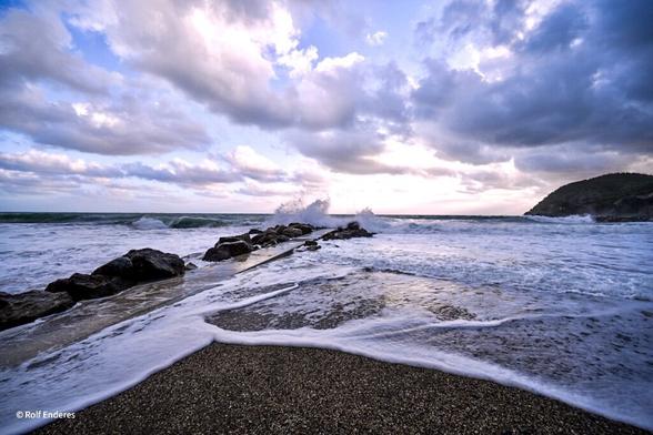 A rocky shoreline with waves crashing against the stones, under a sky filled with dramatic clouds and soft sunlight. The ocean is visible in the background, and a distant hill is seen on the right.