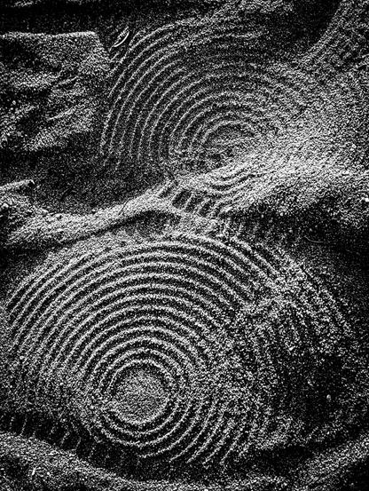 A black and white photograph showing intricate circular and spiral patterns etched into the sand. The patterns resemble those found in a Zen rock garden, meticulously raked to create concentric rings. The two circular designs, one in the upper central portion of the frame and another in the lower central portion, both feature numerous tightly packed, concentric rings.