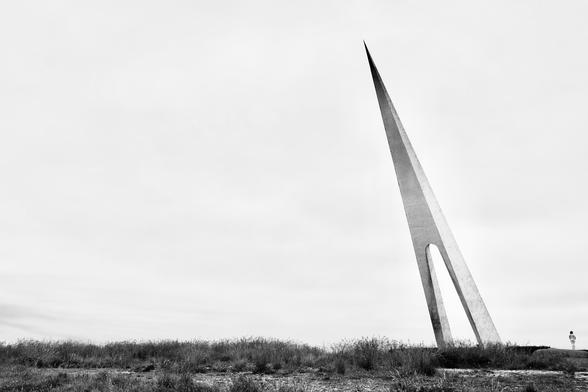 Eine Skulptur ragt gleich einer Kompassnadel spitz aus dem Boden heraus. Zwei säulenartige Betonpfeiler verschmelzen oben zu einer Spitze. Das Gebilde steht im rechten Bildbereich und neigt sich nach links. Die geneigte Haltung nimmt eine rechts daneben stehende Person ebenfalls ein.