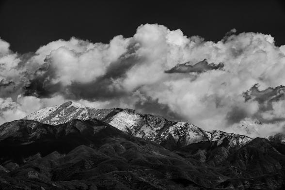 A black and white landscape photograph showing a mountain range with snow-capped peaks. The mountains have rugged, uneven terrain with visible ridges and valleys. The highest peak is prominently snow-covered, while lower elevations show a mix of snow patches and bare rock. Above the mountains, the sky is filled with large, billowing cumulus clouds. The clouds are varied in shape and density, creating a dynamic pattern across the upper portion of the image. The contrast between the bright snow on the peaks and the darker areas of exposed rock is stark, emphasizing the mountain's topography. The foreground shows the lower slopes of the mountains, which appear to be less snow-covered and more densely textured with vegetation or rock formations.