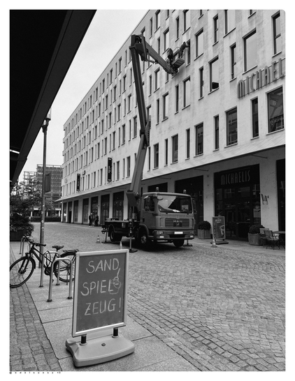 Schwarzweißfotografie: zwischen zwei Gebäuden, eine gepflasterte Ladenstraße, darauf ein Lastkraftwagen mit Hubhebebühne, darauf ein Mann der in der dritten Etage die Fenster reinigt, links ein Aufsteller mit handgeschriebener Werbung: „SAND SPIELZEUG!“ dahinter an einem Fahrradständer ein abgestelltes Fahrrad