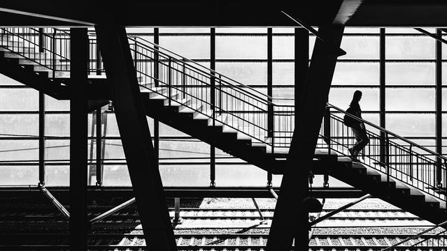 A black and white photo showing a person silhouetted on a staircase inside a modern train station. The scene is framed by angular beams and large window panels that cast a grid of light and shadows over the staircase and train tracks below.