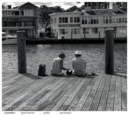 A black and white image of a couple sitting on the edge of a pier in front a river in between two large wooden poles. The woman is looking down at her meal and the man is looking to the left down the river. On the other side of the river bank there is a store or resturant called "Steamboat Inn". There is text in the image stating "Amah's Photography @curious_amadeus@pixelfed.social", "Canon R6 Mark II Sigma Lens 70mm F2.8 June 2024 Mystic, Connecticut United States.