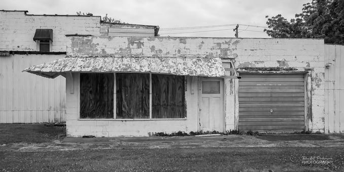 A one-story concrete block building with peeling paint and drooping metal awning over a large triple window and door.