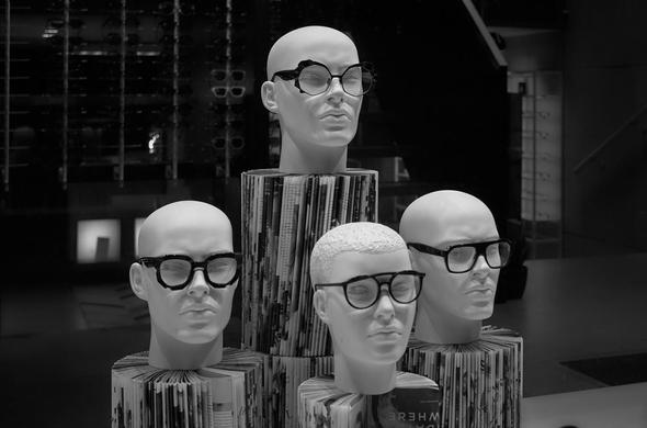 A display featuring four mannequin heads wearing various styles of eyeglasses, arranged on a cylindrical pedestal made of magazines. The image is in black and white.