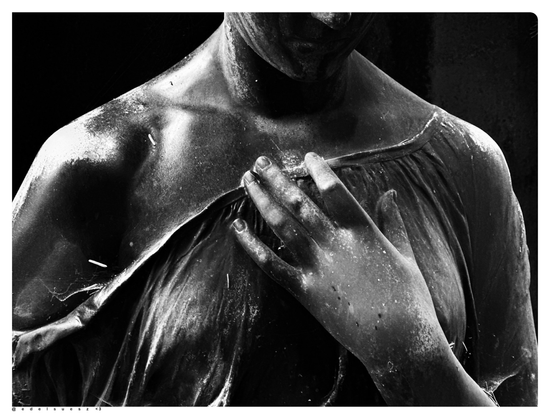 Schwarzweißfotografie: dunkle, nass glänzende Bronzestatue einer Frau, frontal von vorn, sichtbar ist nur der Oberkörper bis zur Nasenspitze, die rechte Schulter ist frei, Sie hält mit ihrer linken Hand das angedeutete Gewand, dunkler Hintergrund