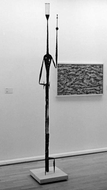 Eeine Statue im Stil von Giacometti, die einen König mit Krone, Schwert und erhobener linker Hand darstellt. Sie befindet sich in einer Kunstgalerie, dahinter ein abstraktes Gemälde.